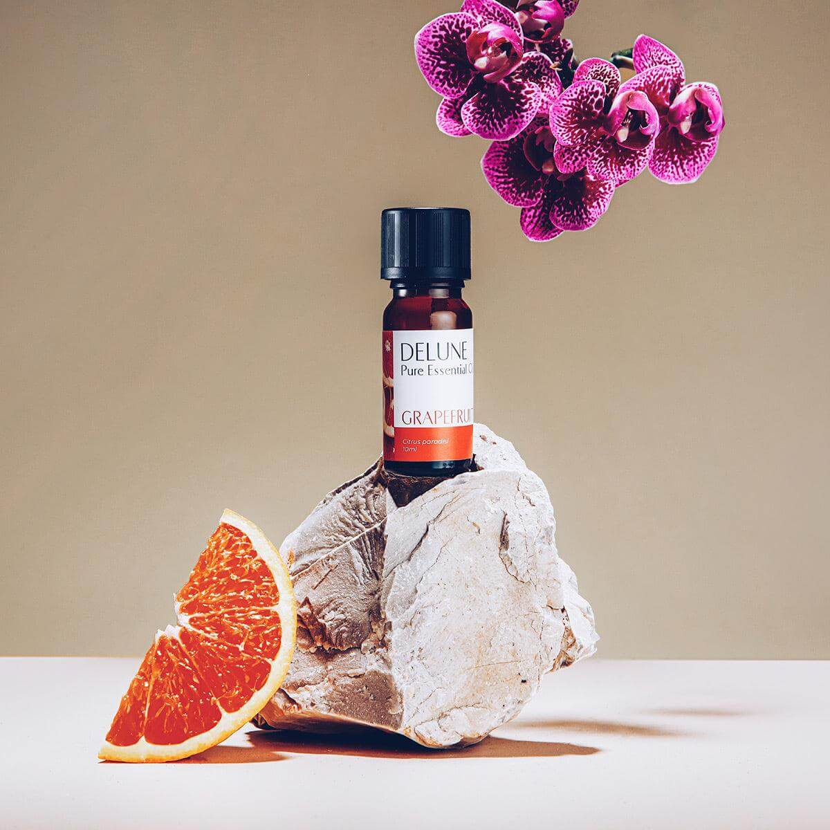 Grapefruit Pure Essential Oil bottle on rock with sliced grapefruit and purple flowers, promoting energy and healthy-looking skin care benefits.