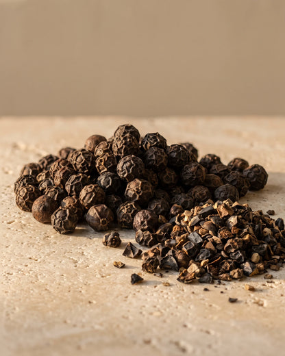 Black Pepper essential oil UAE – Delune Dubai: whole and cracked black peppercorns in raking warm light