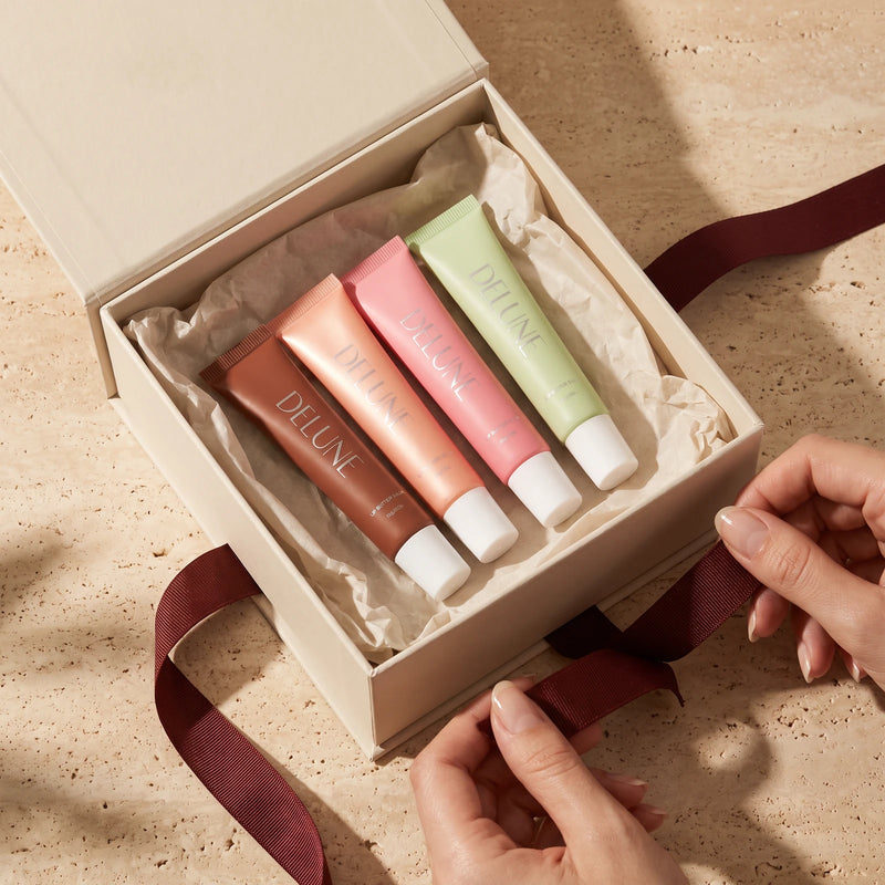 Hands untying an oxblood ribbon from a gift box holding four Lip Butter Balm tubes nested in tissue
