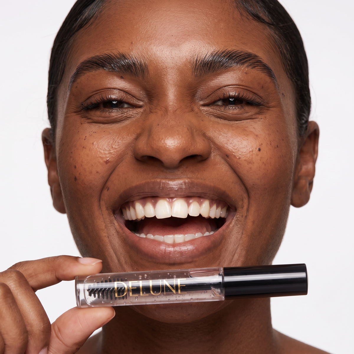 Model laughing with brushed-up brows, holding Delune Dubai Clear Brow Sculpt Gel