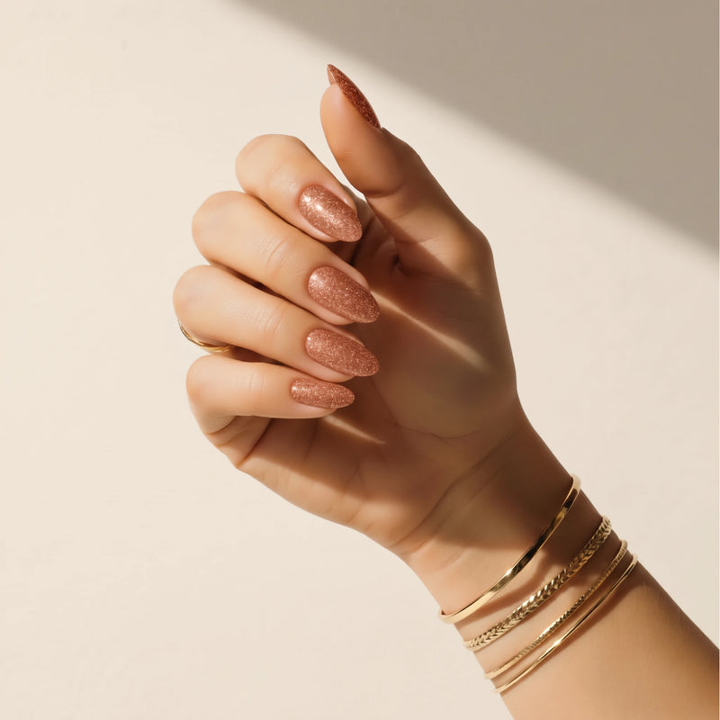 Nail wraps UAE – Hand wearing Delune Dubai Copper Glitter nail wraps with gold bangles against a warm plaster wall