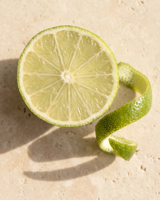 Lime essential oil UAE – Delune Dubai: a halved fresh lime and a curl of lime zest seen from above on warm travertine